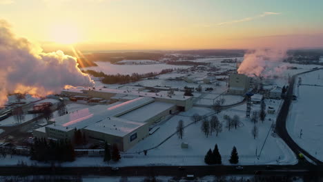 Aerial-pull-back-shot-away-from-a-smoking-factory,-during-a-vibrant-winter-sunset