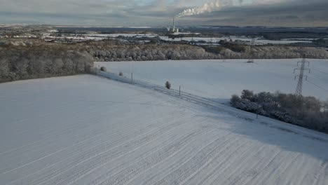 drone-footage-of-snowy-fields,-with-a-power-plant-on-the-horizon