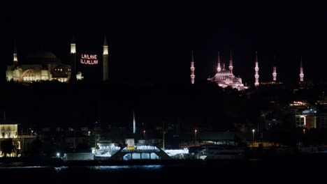Passenger-boat-crossing-the-shore-at-night,-Mosques-with-La-ilahe-illallah-written,-Istanbul,-Turkey