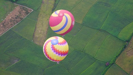 Looking-Down-Over-Pair-Of-Colourful-Hot-Air-Balloons-Flying-Over-Green-Fields-In-Vang-Vieng,-Laos