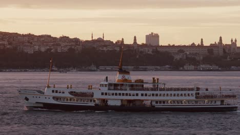 Un-Barco-De-Pasajeros-Navegando-Por-El-Estrecho-Del-Bósforo-Con-Los-Edificios-De-La-Ciudad-De-Estambul-Al-Fondo-Al-Atardecer,-Turquía