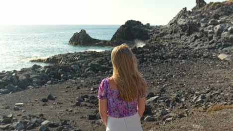 Beautiful-blonde-traveler-adjust-hair-while-admiring-Tenerife-seascape,-back-view