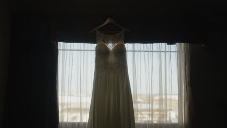 Wunderschönes-Weißes-Hochzeitskleid,-Das-Auf-Einem-Kleiderbügel-Hängt,-Hinterleuchtete-Vorhänge,-Stimmungsvolle,-Szenische-Bewegung-In-Filmischer-Zeitlupe
