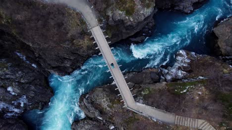 Couple-standing-on-impressive-bridge-over-wild-river-and-waterfall-in-volcanic-landscape,-Aerial-top-down-rotate