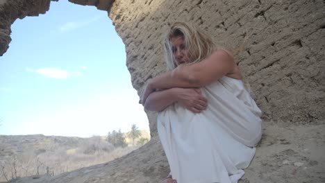 A-woman-in-a-white-dress-huddles-under-an-ancient-ruin-to-shelter-from-the-wind