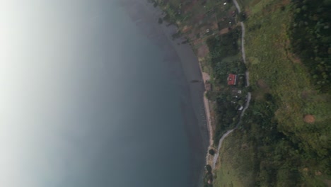 Drone-portrait-view-in-Guatemala-flying-panning-up-from-a-green-mountain-to-a-blue-lake-surrounded-by-green-mountains-and-volcanos-on-a-cloudy-day-in-Atitlan