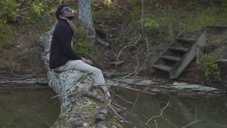 Black-man-in-a-forest-sitting-on-a-tree-trunk-bridge-over-a-creek