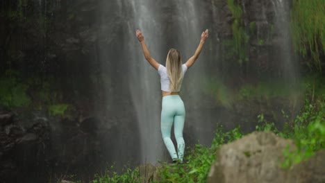 Fit-woman-in-sportive-clothing-reaching-base-of-tropical-waterfall-throwing-hands-up