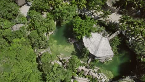 Aerial-view-of-a-cenote-with-traditional-thatched-structures-in-Tulum,-Mexico