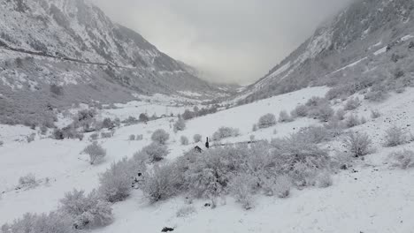 Drohnenaufnahme-Eines-Schneebedeckten-Tals-In-Den-Gebirgigen-Pyrenäen-Spaniens-Mit-Einem-Kleinen-Rustikalen-Haus-Am-Ende-An-Einem-Verschneiten-Tag