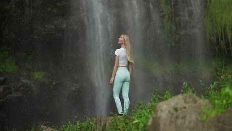 Blond-woman-standing-at-base-of-tropical-waterfall-looking-up,-Agua-D'Alto