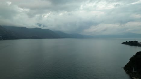 Drone-view-in-Guatemala-panning-a-blue-lake-surrounded-by-green-mountains-and-volcanos-on-a-cloudy-day-in-Atitlan