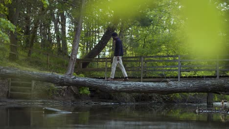 Black-man-standing-and-crossing-a-creek-on-tree-trunk-bridge-in-the-woods---right-to-left