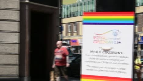 Royal-Borough-Of-Greenwich,-Schwenk-Einer-Ampel-Mit-Schild-Zum-Pride-Month,-Gay-Pride,-Mit-Regenbogen-Fußgängerüberweg-Im-Verschwommenen-Hintergrund,-Tag,-Echtzeit