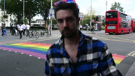 Handsome-man-getting-ready-for-gay-pride-with-pink-sunglasses-in-front-of-rainbow-pedestrian-crossing-in-London-for-pride-month,-eye-level-shot,-static-camera,-day,-real-time