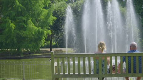 An-old-couple-sitting-on-a-park-bench-in-front-of-a-water-fountain-in-the-summer