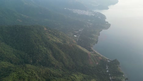 Drone-view-in-Guatemala-panning-down-a-green-mountain-with-a-forest-full-of-trees-and-a-lake-on-a-cloudy-day-in-Atitlan