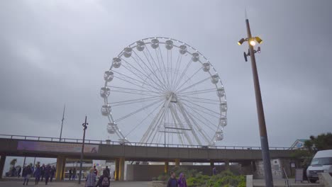A-ferris-wheel-with-flashing-lights-on-an-overcast-afternoon-with-people-walking-in-the-foreground