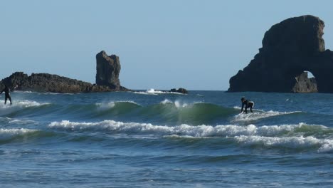 surfers-in-wetsuits-riding-a-wave-across-the-entire-screen-with-rock-ach-in-the-background-pan-right-to-left