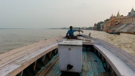 Cinematic-Ancient-Holy-city-Varanasi-India-Ganges-River-man-captain-canal-boat-driver-cruise-Northern-State-people-on-steps-Ghat-Pradesh-Province-landscape-gray-cloudy-fire-smoke-follow-forward