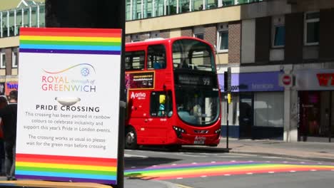 Royal-borough-of-Greenwich,-eye-level-shot-of-traffic-light-with-sign-about-pride-month,-gay-pride,-with-rainbow-pedestrian-crossing-and-iconic-red-double-decker-bus-in-blurred-background,-day