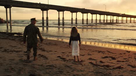 Follow-shot-behind-military-male---young-female-in-flowing-white-dress-walking-on-sunset-glow-sandy-beach-with-waves-lapping-in