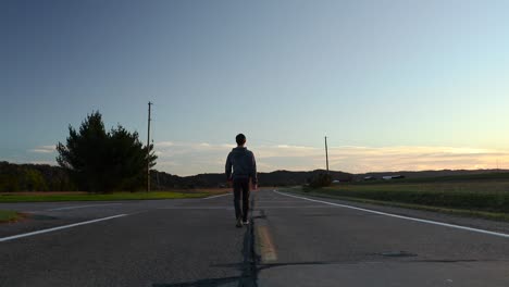 A-man-walks-slowly-away-into-the-distance-down-the-middle-of-a-rural-cracked-asphalt-road-at-dusk