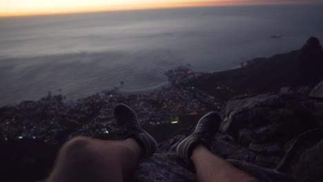 View-of-Cape-Town-and-the-Atlantic-Ocean-at-night-from-Table-Mountain-above-a-Mans-feet,-in-Slow-Motion