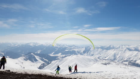 Paraglider-pilot-with-yellow-sail-runs,-launches-and-flies-toward-spectacular-snow-capped-Caucasus-mountain-range,-behind-pan