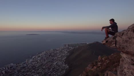 Man-sitting-on-the-edge-of-the-cliff,-enjoying-the-view-of-Cape-Town,-South-Africa-from-Lions-Head-during-Sunset,-in-Slow-Motion