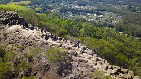 Aerial-tracking-drone-shot-of-a-group-of-people-hiking-on-a-rocky-mountain,-on-a-sunny-day,-in-Glass-house-mountains-national-park,-in-Queensland,-Australia