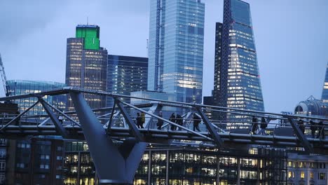 Menschen-Gehen-Durch-Die-Millennium-Bridge-In-London,-England,-Vereinigtes-Königreich-Mit-Beleuchteten-Wolkenkratzern-Im-Hintergrund