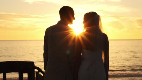 Rear-partial-silhouette-panning-shot-across-happy-couple-bride---groom-on-paradise-beach-kiss-as-sunset-reveals-between-them