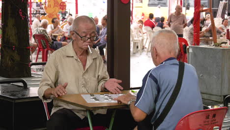 Dos-Ancianos-Encienden-Un-Cigarrillo-Mientras-Juegan-Ajedrez-Chino-En-Chinatown,-Singapur