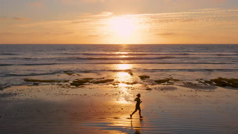 Aerial-passing-by-the-profile-of-a-woman-running-along-the-beach-at-low-tide-at-sunset