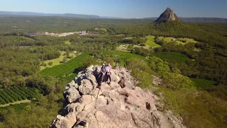 Aerial-drone-shot-away-from-a-couple,-man-and-woman-hiking-on-a-rocky-mountain,-on-a-sunny-day,-in-Glass-house-mountains-national-park,-in-Queensland,-Australia