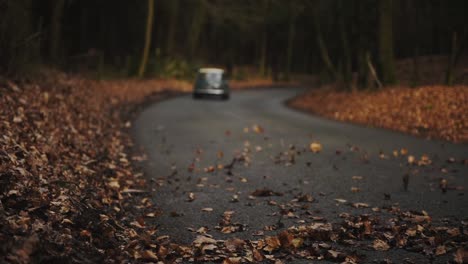 Old-classic-car-mini-driving-on-a-road,-speeding-past-fallen-autumn-leaves-in-a-forest,-England