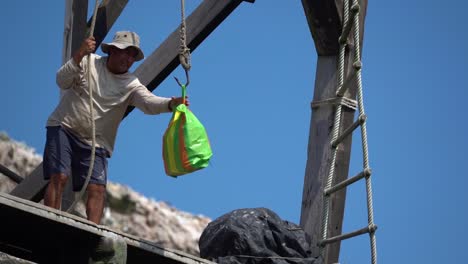 Peruvian-man-pulling-up-a-colorful-plastic-bag-on-a-hook-in-harbor,-slow-motion