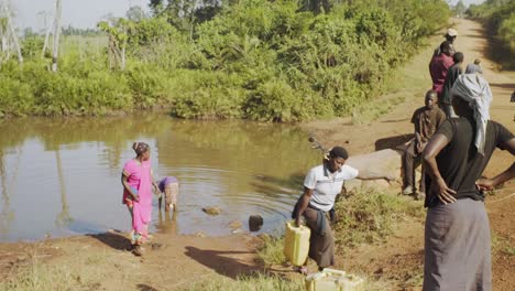 A-villager-woman-carrying-a-full-water-container-up-towards-the-dirt-road-while-others-are-waiting