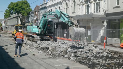 wide-shot-Workman-sweeping-as-digger-breaks-road-concrete-to-replace-tram-lines,-bulldozer-behind