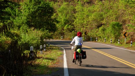 A-lone-female-cyclist-mounts-and-then-rides-along-a-road-near-Paknai's-Fisherman-Village-in-the-Na-Muen-District,-Nan,-Thailand