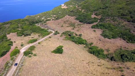 Drone-aerial-shot,-lowering-down-and-following-a-black-car-in-the-coast-road-on-a-mediterranean-landscape-in-Sardiniai-Italy