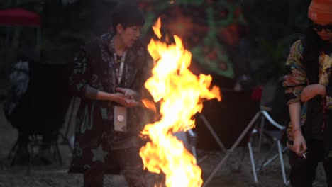 Asian-male-and-female-hanging-around-long-orange-flames-coming-from-fire-pit-in-outdoor-setting,-filmed-in-handheld-style-as-medium-close-up-in-slow-motion