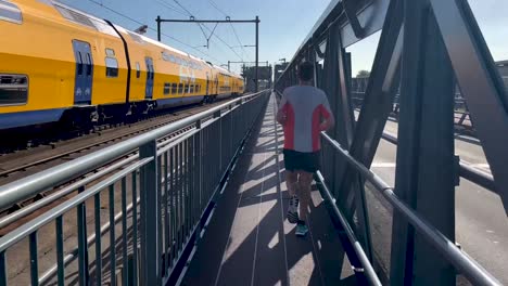 Passing-Dutch-train-following-of-male-trail-runner-seen-from-behind-on-steel-draw-bridge-along-train-tracks