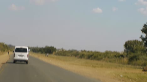 Lieferwagen-Und-Lastwagen-Rasen-über-Die-Asphaltstraße,-Während-Kleine-Kinder-Auf-Einem-Unbefestigten-Weg-In-Afrika-Laufen