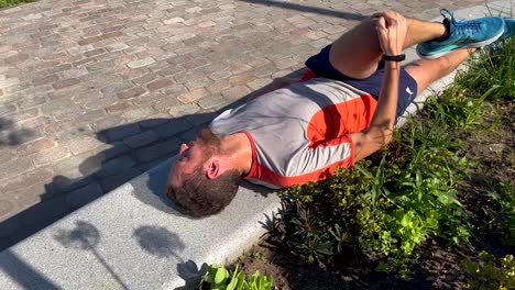 Stretch-and-breathing-exercises-cool-down-after-training-run-session-of-a-male-trail-runner-laying-on-public-bench-beside-vegetation