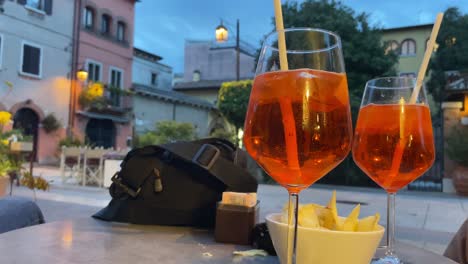 Statische-Ansicht-Vom-Tisch-Mit-Drei-Freunden,-Die-Bei-Sonnenuntergang-In-Der-Straße-Eines-Traditionellen-Dorfes-Typisch-Italienischen-Aperol-Spritz-Trinken
