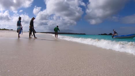 Touristen-Mit-Schnorchelausrüstung-Laufen-An-Einem-Strand-Mit-Weißem-Sand-Und-Türkisfarbenem-Wasser-Der-Galapagos-Inseln-Spazieren,-Während-Ein-Boot-Näher-An-Die-Küste-Herankommt,-Um-Sie-Abzuholen
