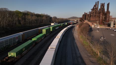 An-aerial-view-of-cargo-trains-in-Bethlehem,-Pennsylvania-on-a-sunny-day,-with-the-Bethlehem-Steel-plant-in-the-background