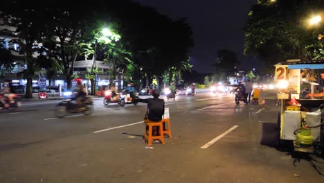 man-sitting-next-to-the-road,-waving-money-to-exchange-to-cars-driving-by,-motor-scooters-passing,-city-lights-in-a-dark-street-in-surabaya,-Indonesia